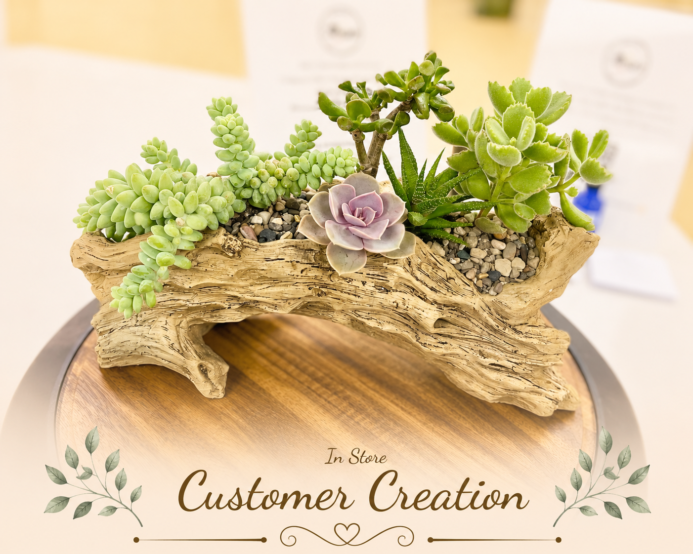 A decorative succulent arrangement in a natural driftwood planter, displayed on a wooden pedestal with a blurred indoor background and the text 'In Store Customer Creation' overlaid at the bottom.