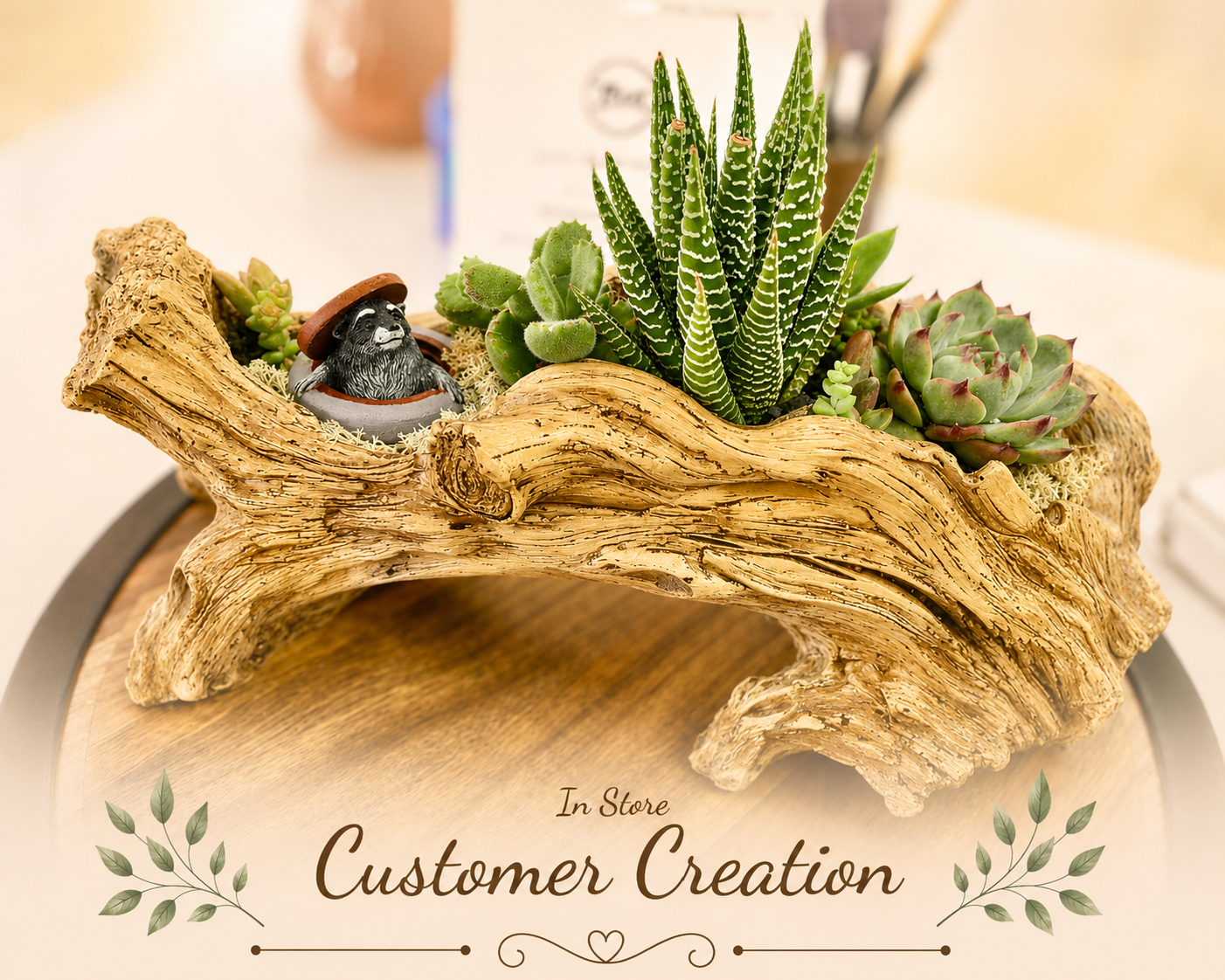 A decorative in-store customer creation planter made from a natural driftwood log, filled with small green succulents and a small potted bird figurine, displayed on a wooden surface with a blurred store background.