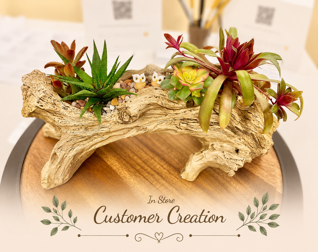A decorative arrangement of small potted succulents and air plants set in a hollow driftwood log, displayed on a round wooden tray. The background is softly blurred, and a decorative 'In Store Customer Creation' watermark overlay is present at the bottom.