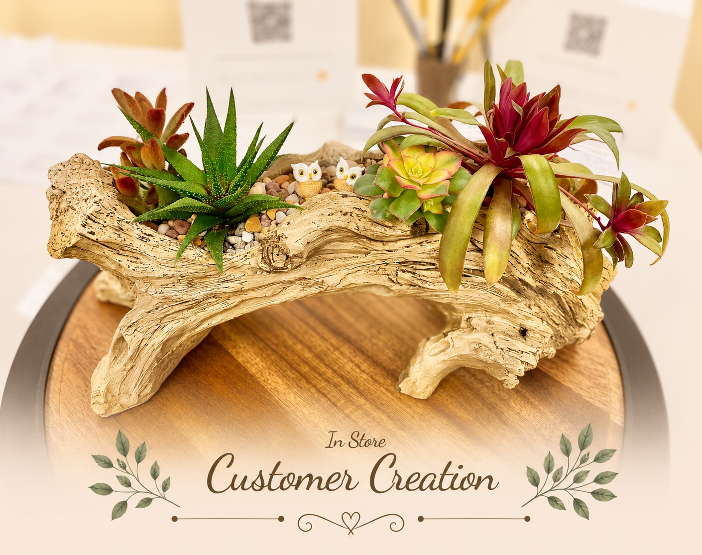 A decorative arrangement of small potted succulents and air plants set in a hollow driftwood log, displayed on a round wooden tray. The background is softly blurred, and a decorative 'In Store Customer Creation' watermark overlay is present at the bottom.