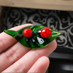 A hand holding a green leaf-shaped decorative piece with two red ladybugs, shown in front of a softly blurred indoor background with small figurines on a shelf.