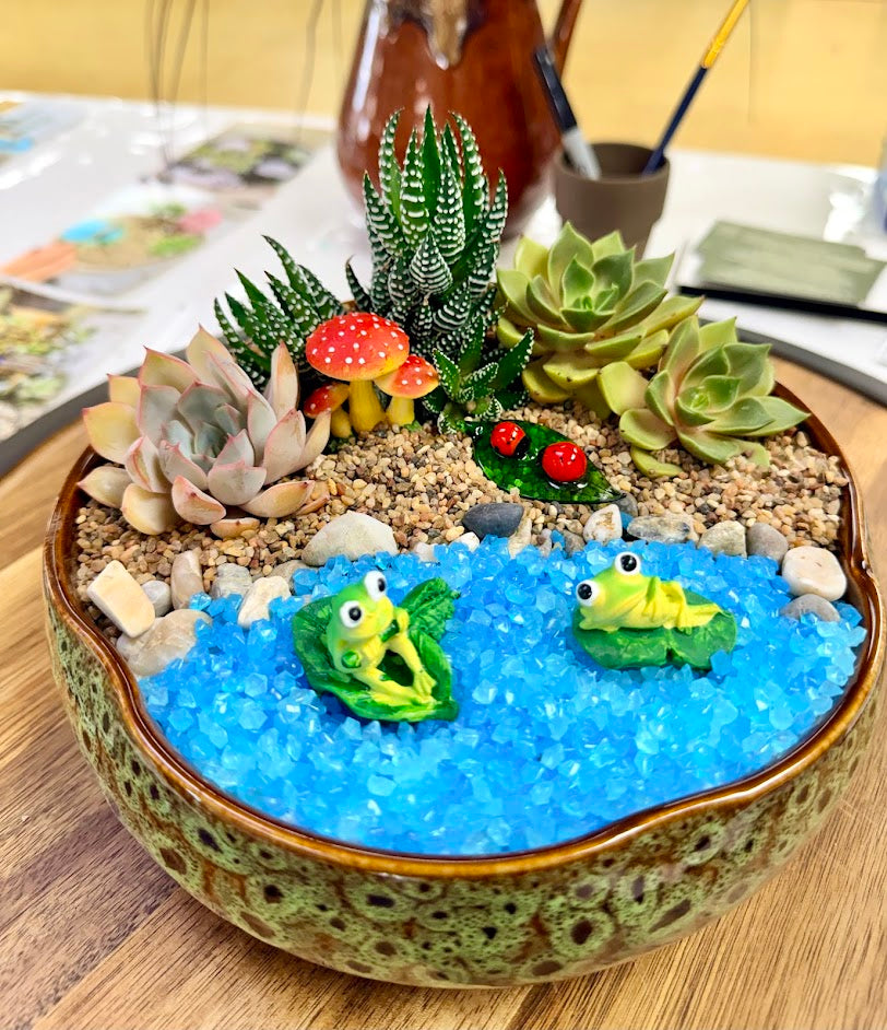 A decorative succulent and moss garden arranged in a shallow ceramic bowl filled with bright blue gravel. The garden includes small green succulents, a striped succulent, red-and-white mushrooms, a small green leafy plant, and two small green frog figurines on top of the blue gravel.