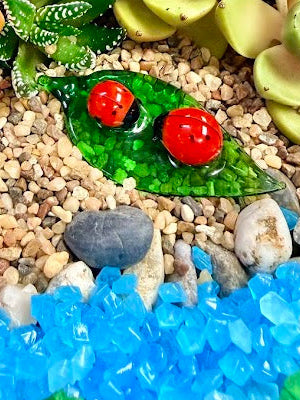 close up of a handmade glass leaf with two red ladybugs on it. it is laid on a small gravel beach in a fairy garden