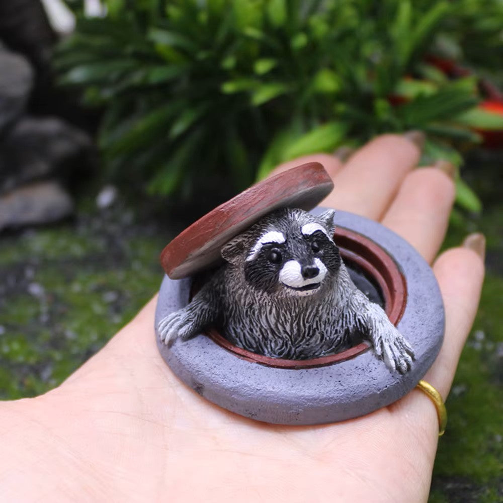 Hand holding a small sculpture of a raccoon in a manhole with a natural background